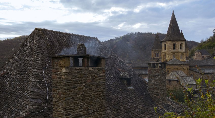 Conques in the Global World (concluded) Stone rooftops with chimneys, a tower in the background, and hills under a cloudy sky.