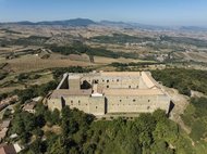 Aerial view of a massive stone fortress amidst lush greenery, with expansive fields and distant mountains in the background.